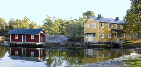 Två hus invid vattnet med skog i bakgrunden. Huset till höger är gult och i två våningar, det till vänster är rött och i en våning.